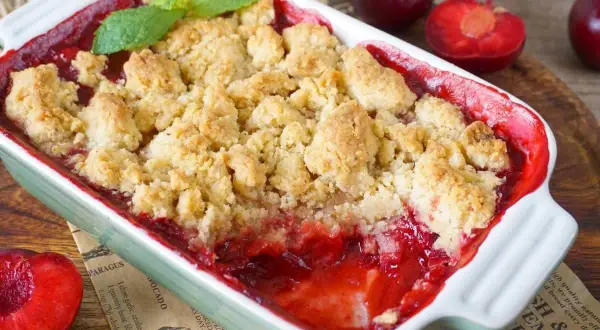 Ein warmer pflaumen crumble mit knuspriger Streuseldecke und frischer Minze. Daneben liegen saftige Pflaumen.