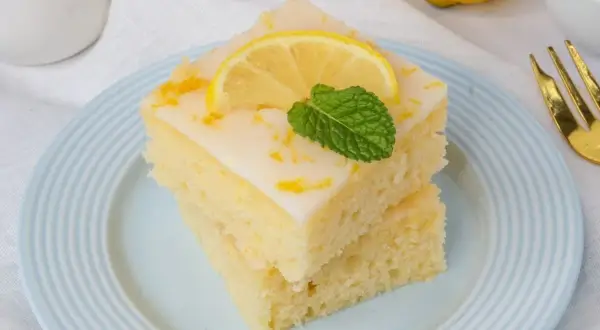 Zwei Stücke zitronenkuchen mit joghurt, garniert mit Zitronenscheibe und Minze, auf blau-weiß gestreiftem Teller.