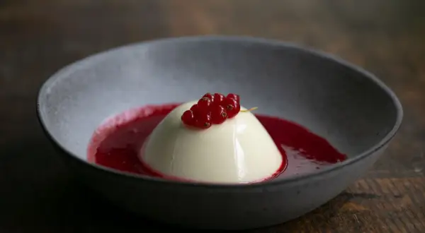 Cremige Panna Cotta mit roter Beerensauce und frischen Johannisbeeren in einer grauen Schale auf Holzhintergrund.