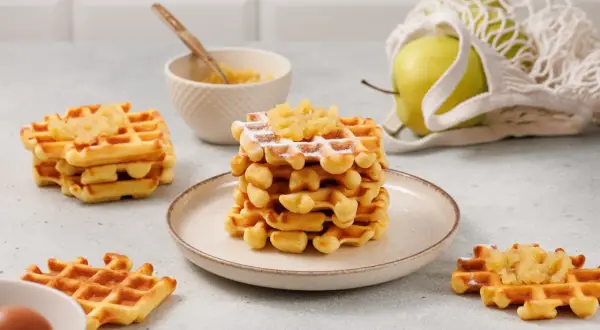 Gestapelte Waffeln mit Apfelmus bestreut, daneben ein Apfel im Netzbeutel und eine Schüssel mit Apfelmus.
