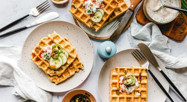 Drei Teller mit veganen Waffeln, garniert mit Gurkenscheiben, Zwiebeln und einer cremigen Sauce. Dazu ein Schälchen mit eingelegten Jalapeños.