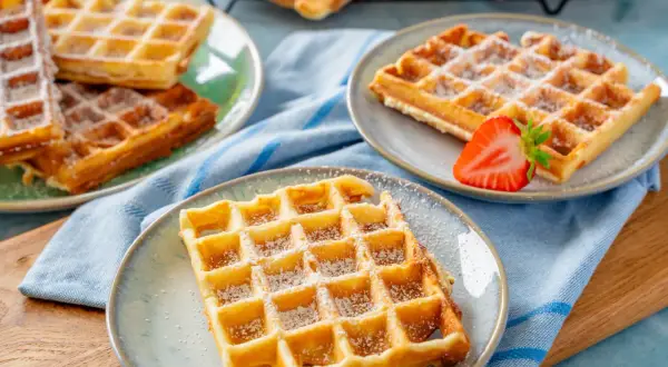 Goldbraune Waffeln, bestäubt mit Puderzucker, warten auf ein leckeres Waffeln Rezept. Eine Erdbeere garniert eine Waffel.