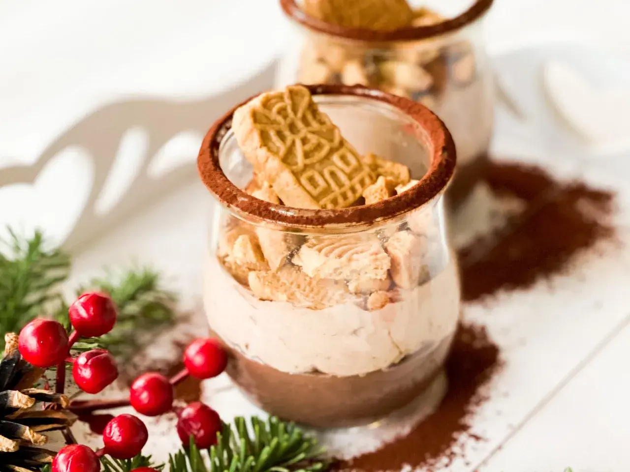 Zwei Gläser mit cremiger Spekulatius-Creme, garniert mit Spekulatius-Stücken und Kakaopulver. Weihnachtliche Deko mit Tannenzweigen und roten Beeren.