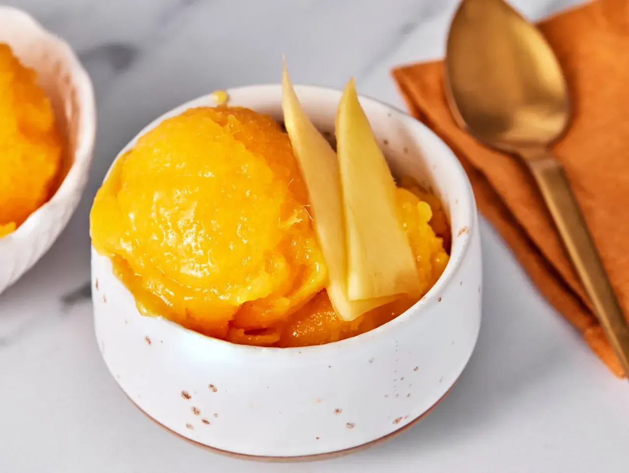 Erfrischendes Mango-Sorbet in einer Schale, garniert mit Mangostücken. Daneben liegt ein goldener Löffel auf einem orangen Tuch.