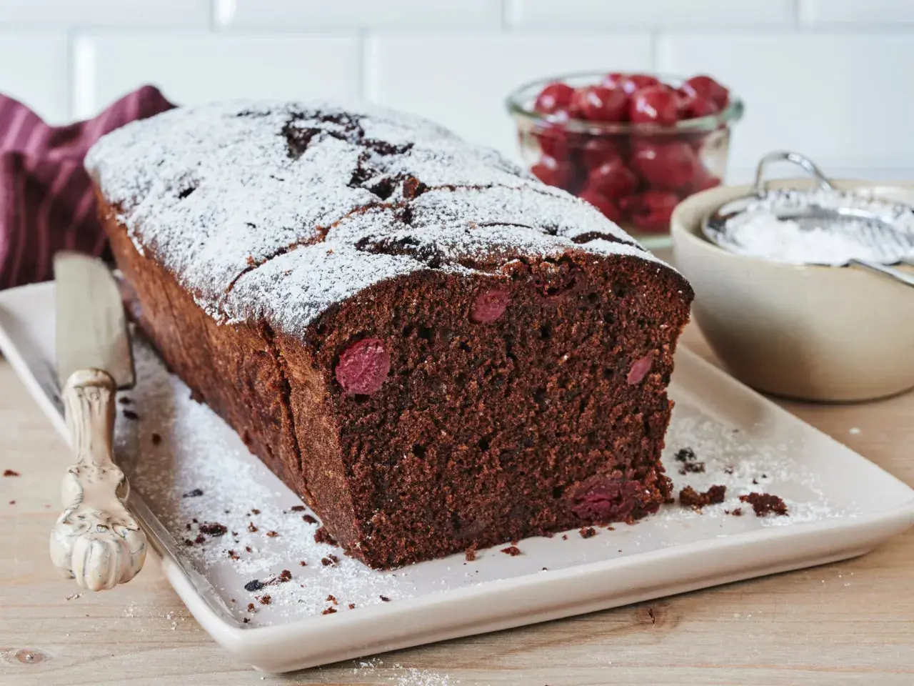 Saftiger schoko kirsch kuchen, bestäubt mit Puderzucker. Daneben eine Schale mit Kirschen und ein Sieb.