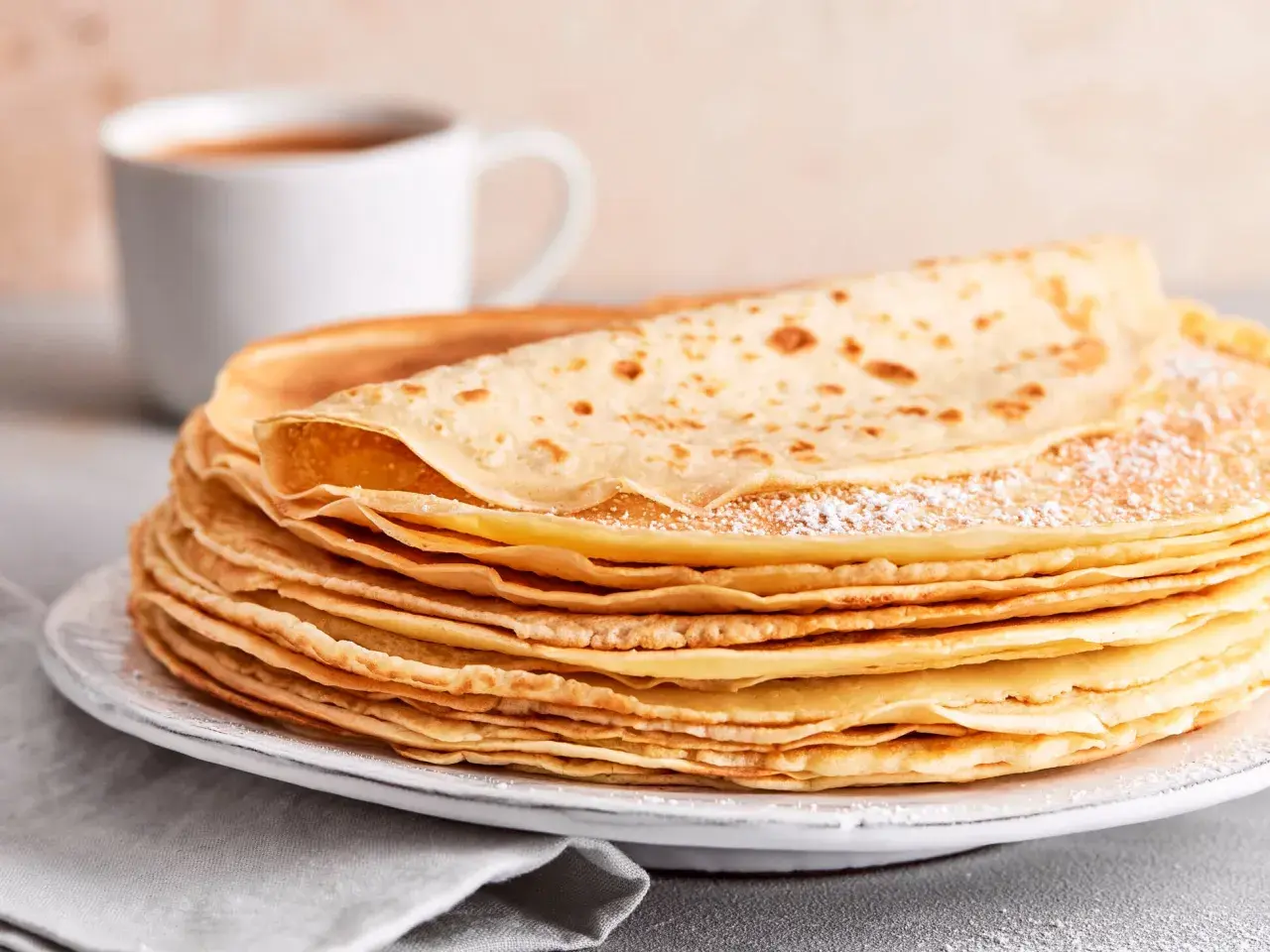 Ein Stapel goldbrauner Pfannkuchen, bestäubt mit Puderzucker, wartet auf ein leckeres Pfannkuchen Rezept. Daneben eine Tasse Kaffee.