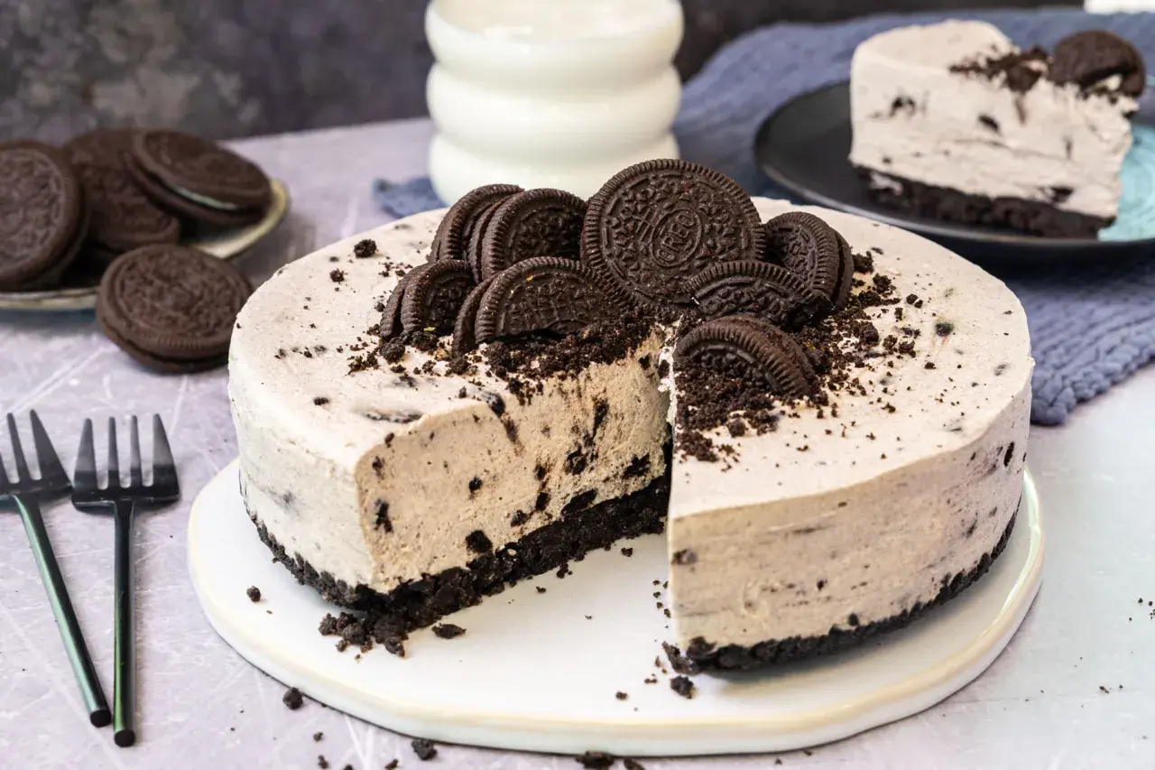 Ein köstlicher Oreo Kuchen, dekoriert mit ganzen und zerbrochenen Keksen, wartet darauf, angeschnitten zu werden. Daneben liegen Gabeln und ein Glas Milch.