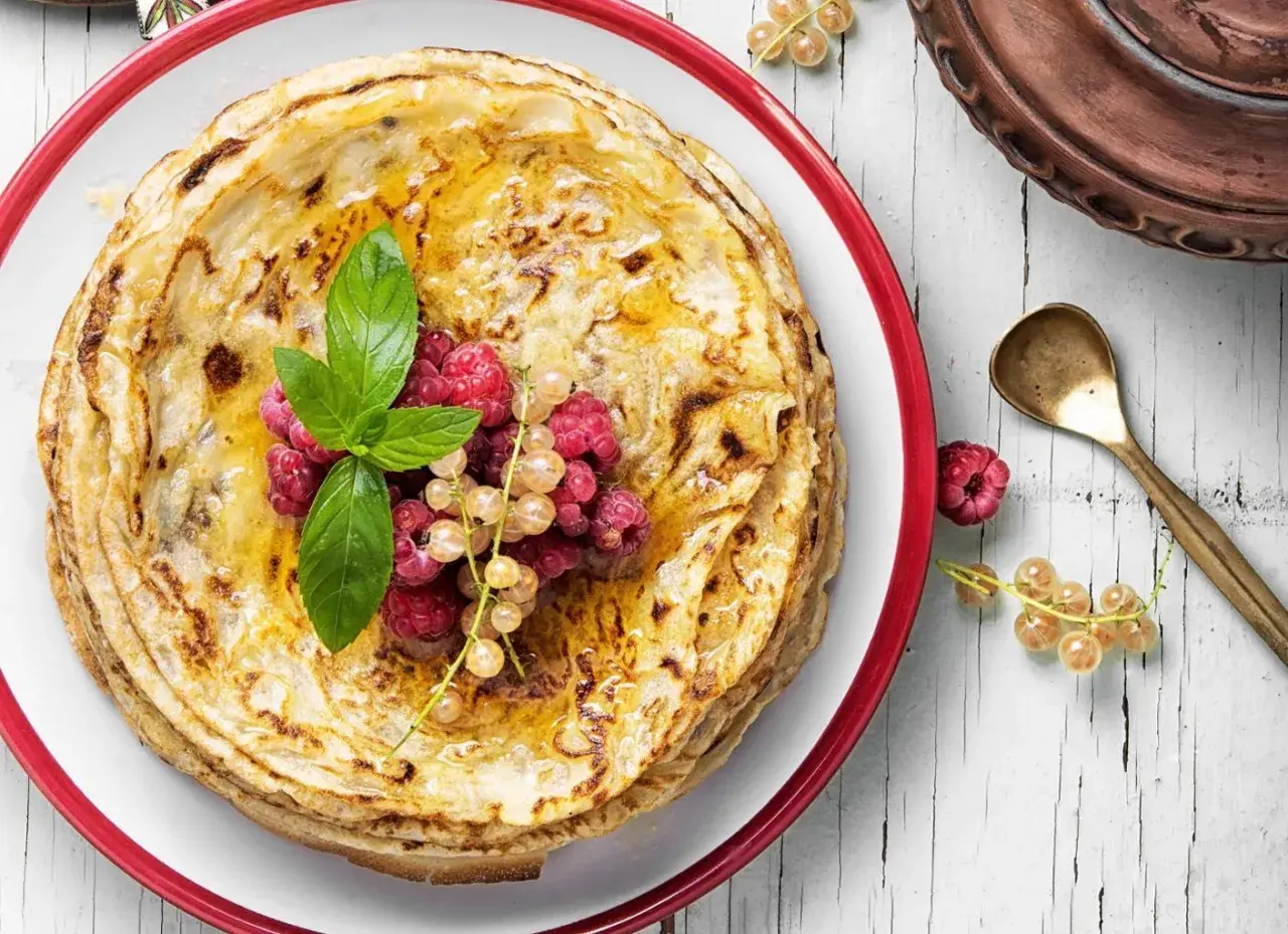 Ein Stapel goldbrauner Pfannkuchen, garniert mit Beeren und Minze. Der Pfannkuchenteig ist perfekt gebacken und mit Honig beträufelt.