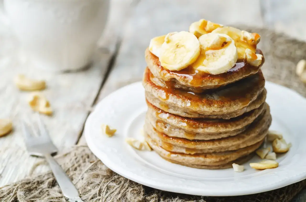 Ein Stapel goldbrauner Bananenpfannkuchen, beträufelt mit Sirup und garniert mit Bananenscheiben und Cashewkernen.