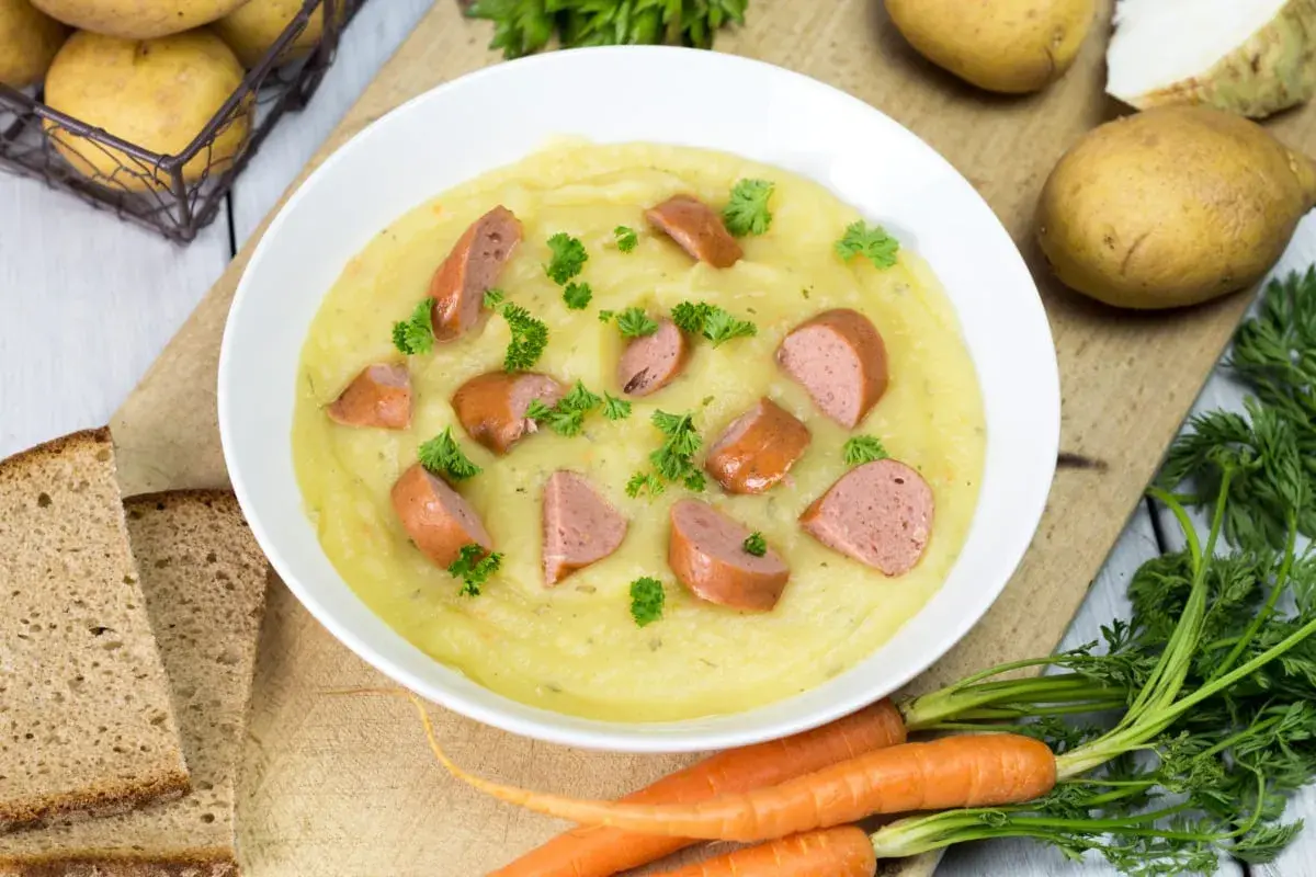 Cremige Kartoffelsuppe mit Würstchenstücken und Petersilie, zubereitet im Thermomix. Dazu gibt es Brot und frische Karotten.