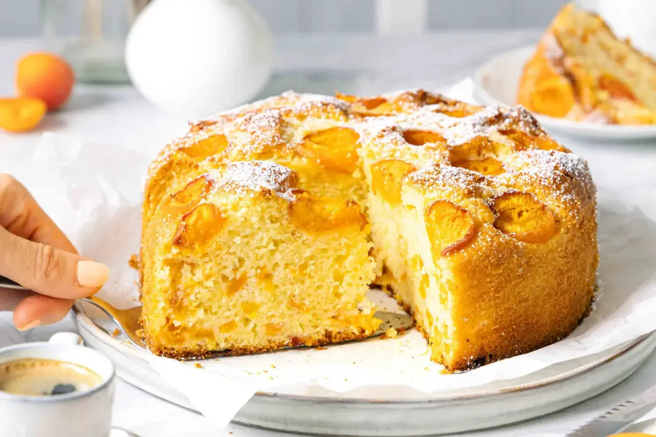 Ein saftiger Aprikosenkuchen, bestäubt mit Puderzucker, wird angeschnitten. Ein Stück davon könnte der weltbester Aprikosenkuchen sein.