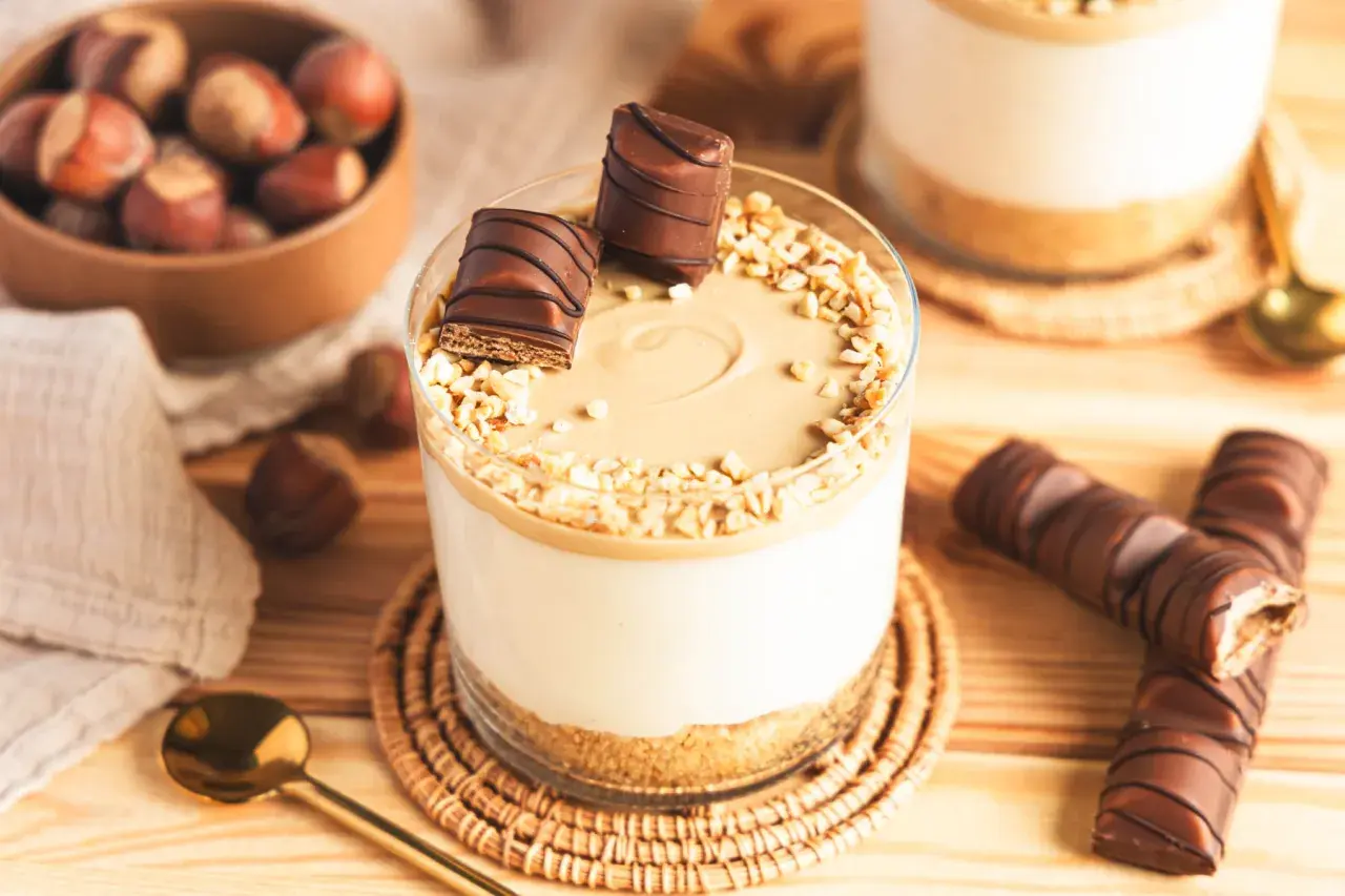 Ein Glas mit cremiger Kinder Bueno Creme, garniert mit Nüssen und Schokostückchen. Daneben liegen ganze Kinder Bueno Riegel.