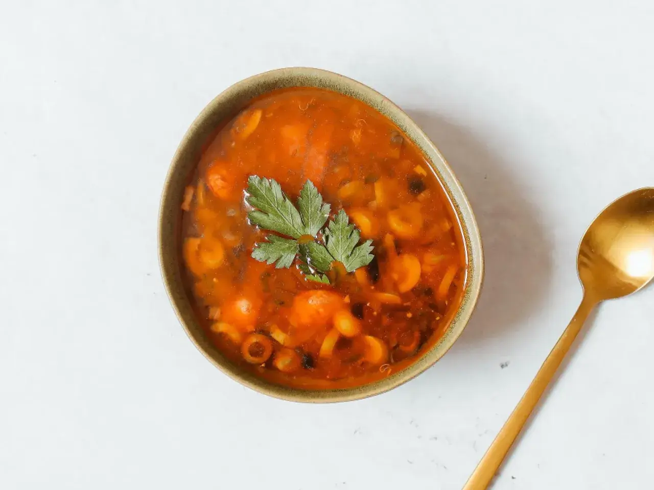 Herzhaftes Soljanka Rezept: Eine Schüssel voller würziger Suppe mit Oliven, Kapern und Kräutern, daneben ein goldener Löffel.