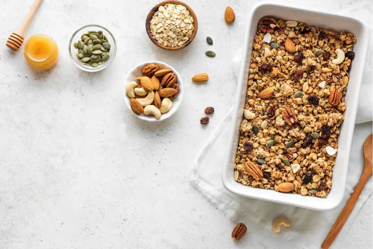Zutaten für ein leckeres baked oats rezept: Haferflocken, Nüsse, Kürbiskerne, Rosinen und Honig in Schalen, daneben eine Auflaufform mit fertigem Granola.