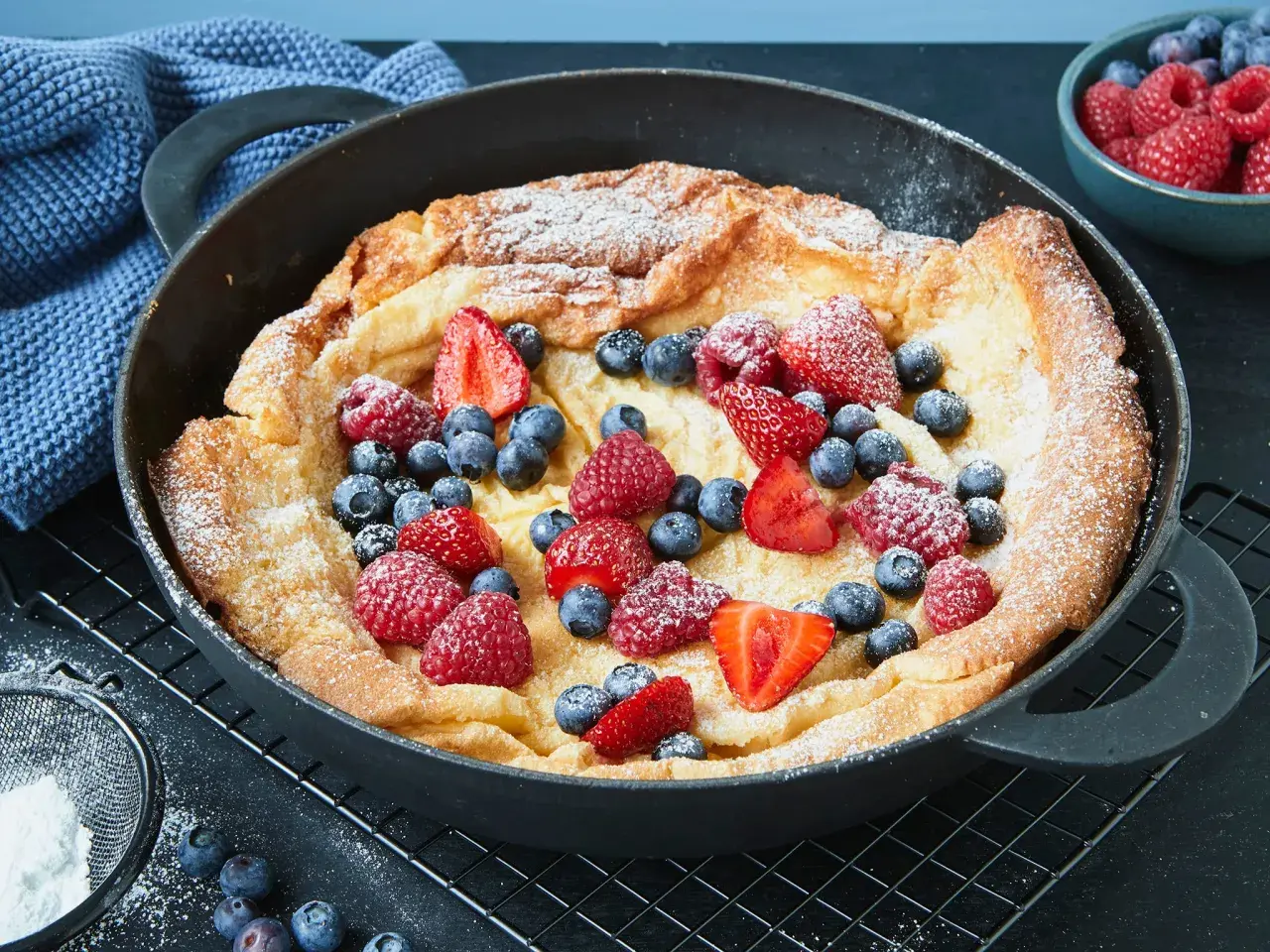 Ein goldbrauner Dutch Baby mit Puderzucker und frischen Beeren wie Erdbeeren, Himbeeren und Blaubeeren in einer Gusseisenpfanne.