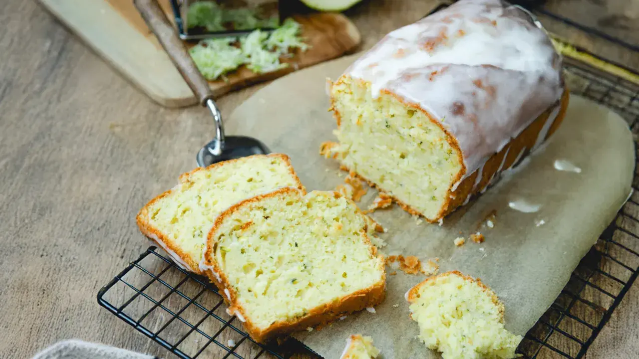 Saftiger Zucchini Kuchen mit Zuckerguss, frisch angeschnitten auf einem Gitter. Daneben liegen geriebene Zucchini und ein Hobel.