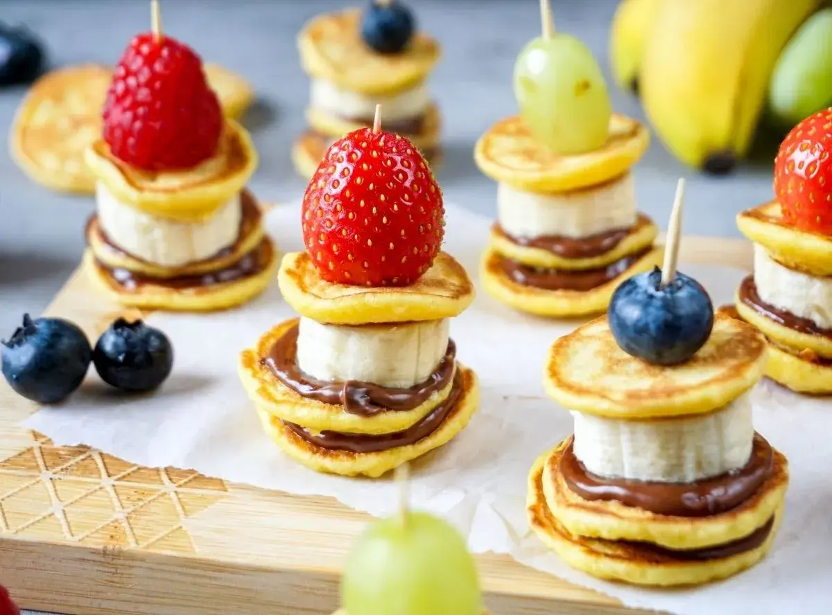 Kleine, gestapelte Baby-Pfannkuchen mit Banane und Schokoaufstrich, garniert mit Beeren und Trauben. Ein süßer Genuss!
