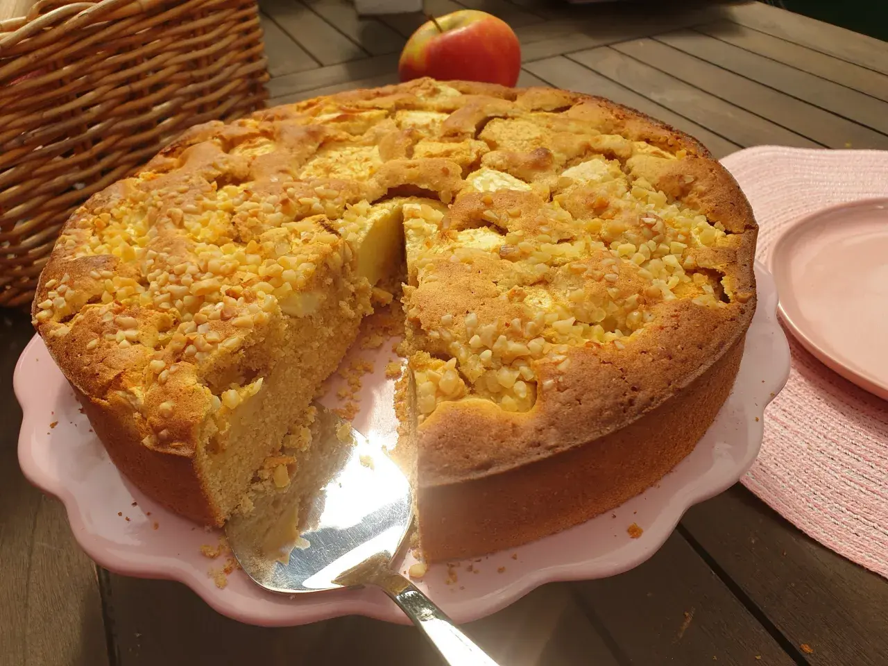 Ein goldbrauner Apfelkuchen ohne Zucker, bestreut mit Nüssen, wird auf einem rosa Teller präsentiert. Daneben liegt ein roter Apfel.