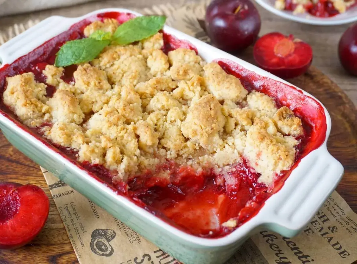 Ein warmer pflaumen crumble mit knuspriger Streuseldecke und frischer Minze. Daneben liegen saftige Pflaumen.