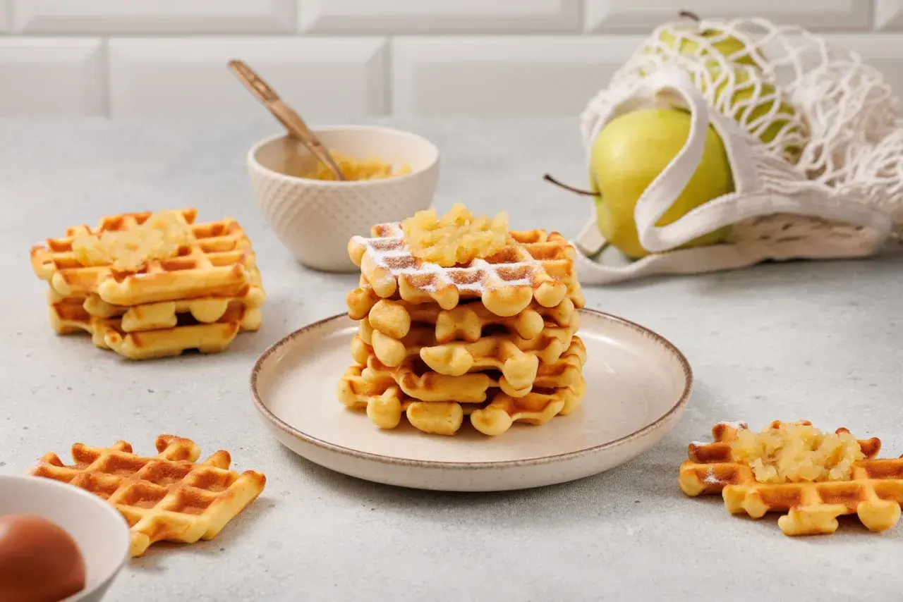 Gestapelte Waffeln mit Apfelmus bestreut, daneben ein Apfel im Netzbeutel und eine Schüssel mit Apfelmus.