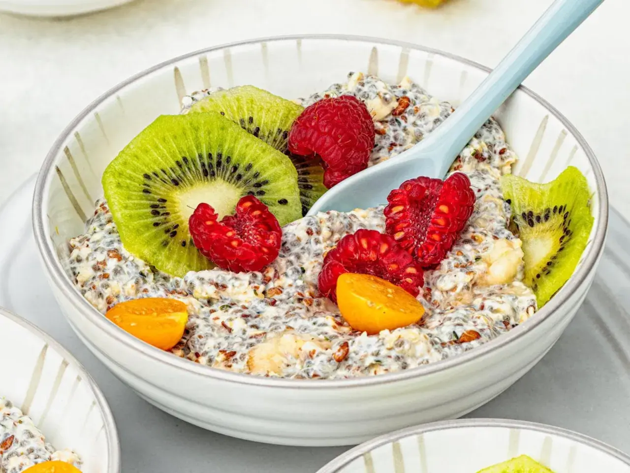 Schüssel mit Overnight Oats, garniert mit Kiwi, Himbeeren und Physalis. Ein Löffel liegt darin.