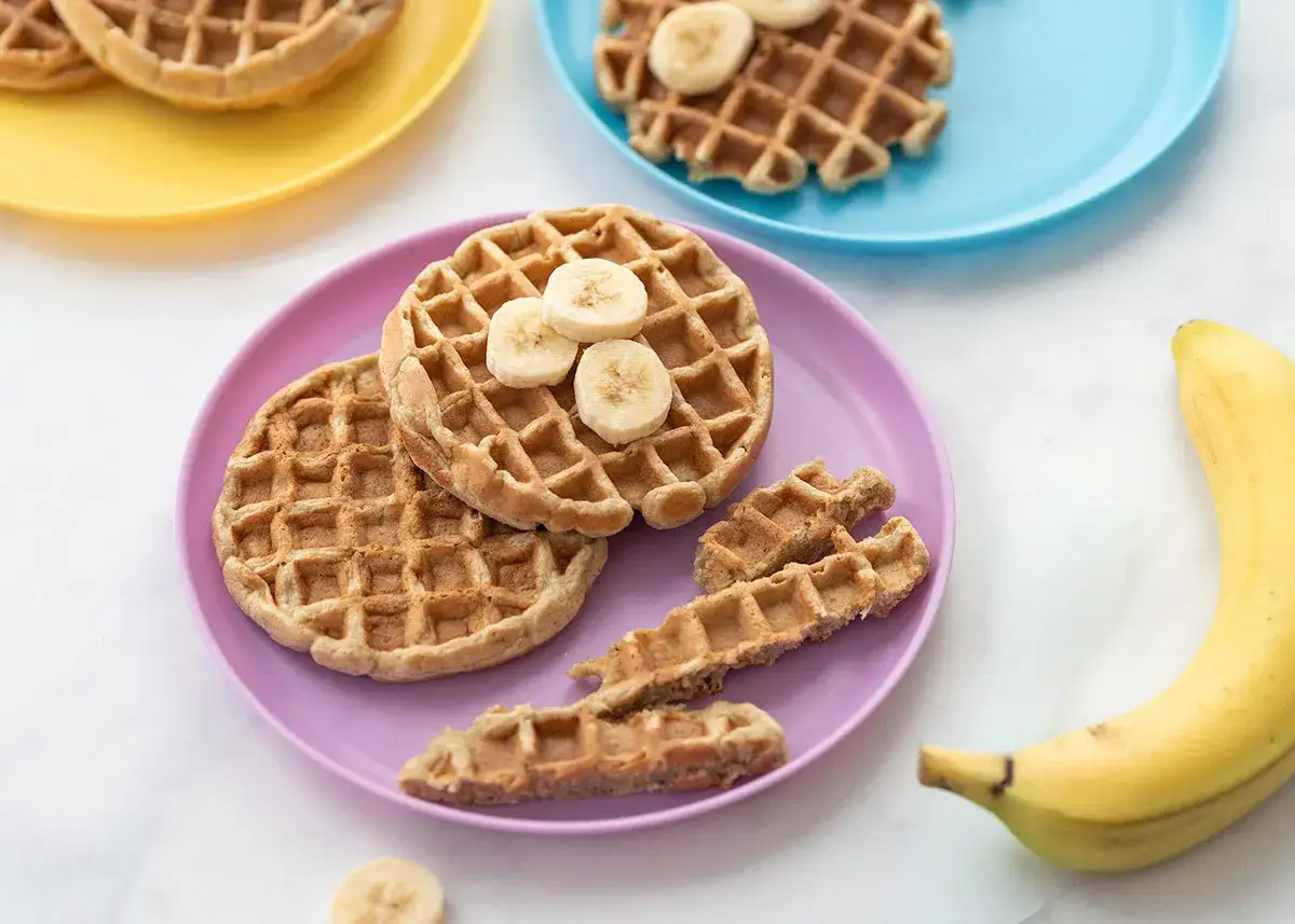 Kleine, runde Baby Waffeln mit Bananenscheiben auf einem lila Teller. Daneben liegen knusprige Waffelst&auml;bchen und eine Banane.