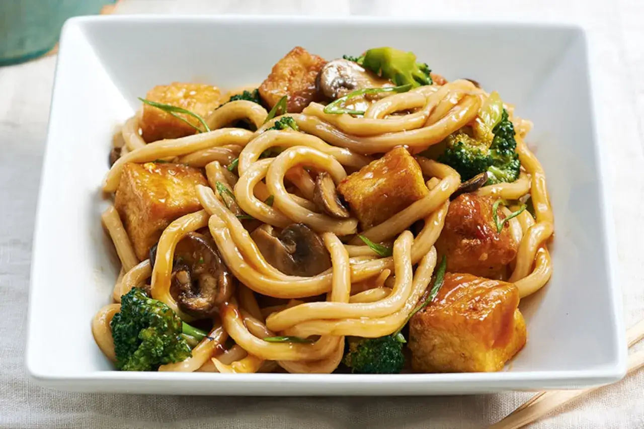 Herzhafte udon nudeln mit tofu, Brokkoli und Pilzen in einer k&ouml;stlichen Sauce, serviert in einer wei&szlig;en Sch&uuml;ssel.