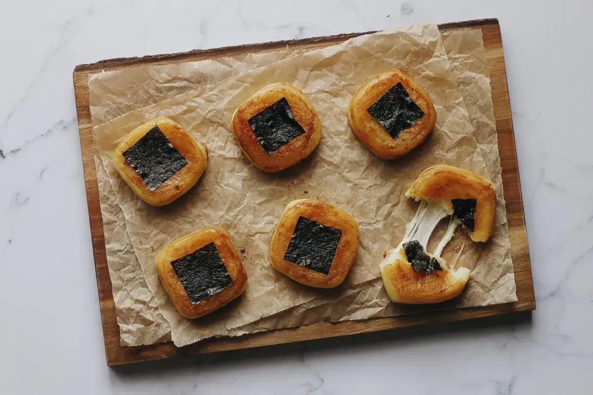 Sechs quadratische, goldbraune Mochi mit schwarzem Nori-Topping liegen auf Backpapier. Einer ist aufgerissen und zeigt z&auml;hfl&uuml;ssigen K&auml;se. Perfekt zum Mochi selber machen!