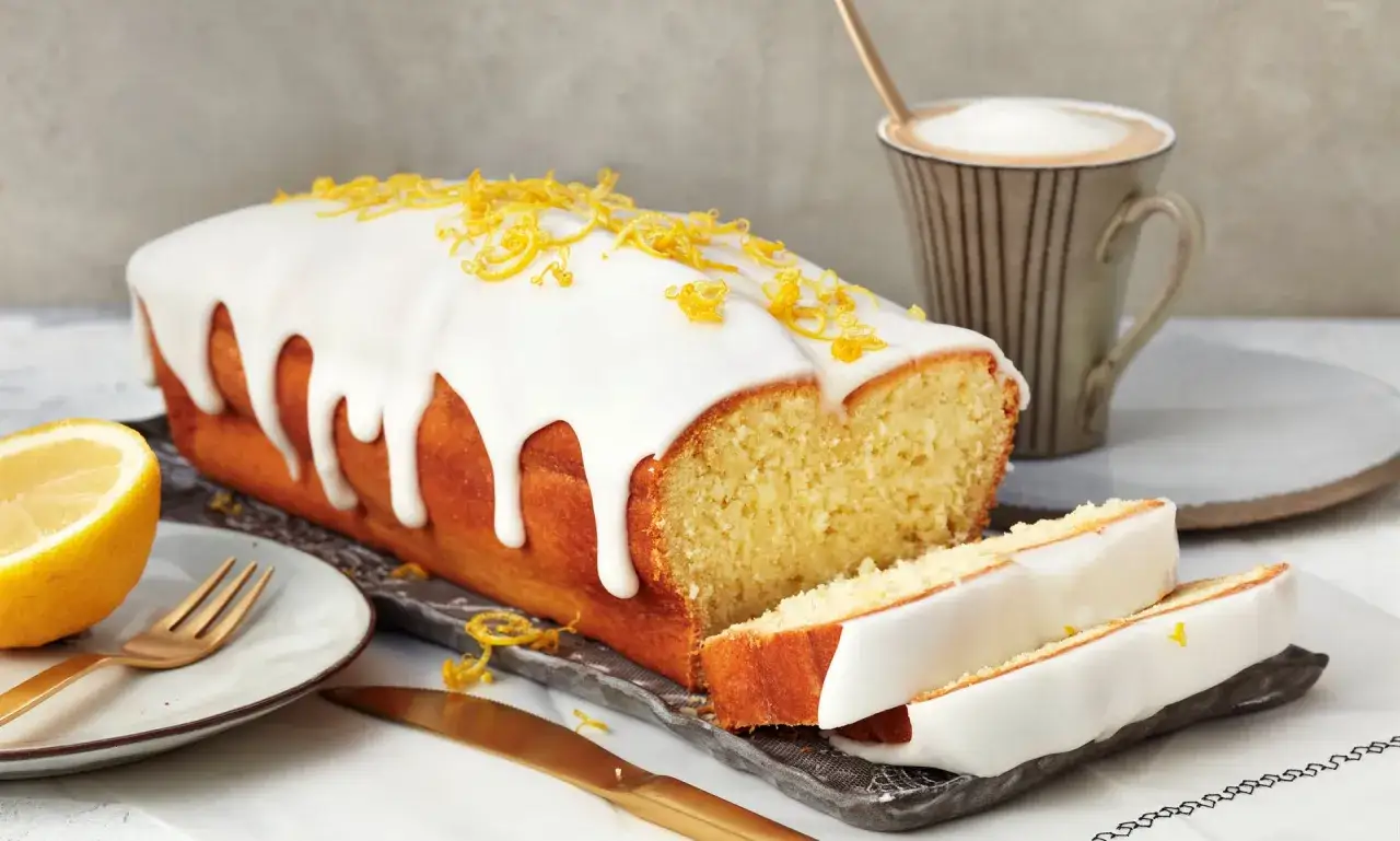 Saftiger zitronenkuchen mit Joghurt, &uuml;berzogen mit wei&szlig;er Glasur und garniert mit Zitronenschale. Daneben eine Tasse Kaffee.