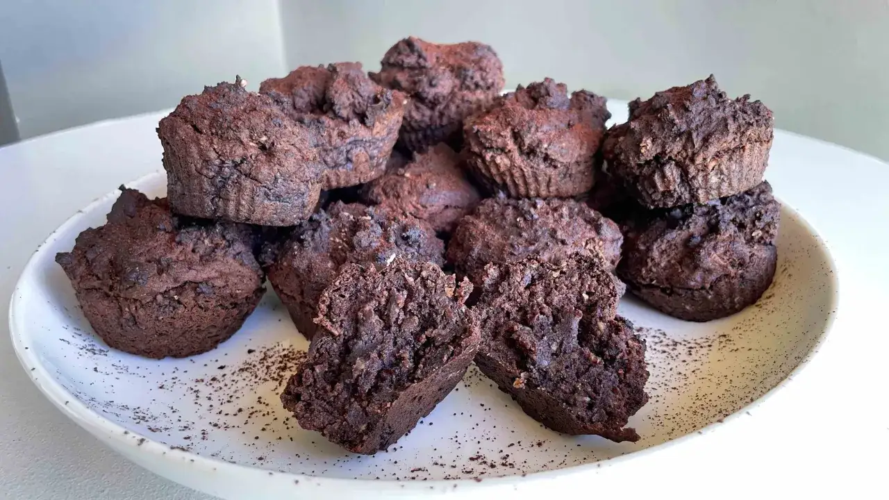 Schokoladen-Muffins mit Proteinpulver, auf einem Teller mit Kakaopulver bestreut. Einer ist aufgeschnitten und zeigt die saftige Textur.