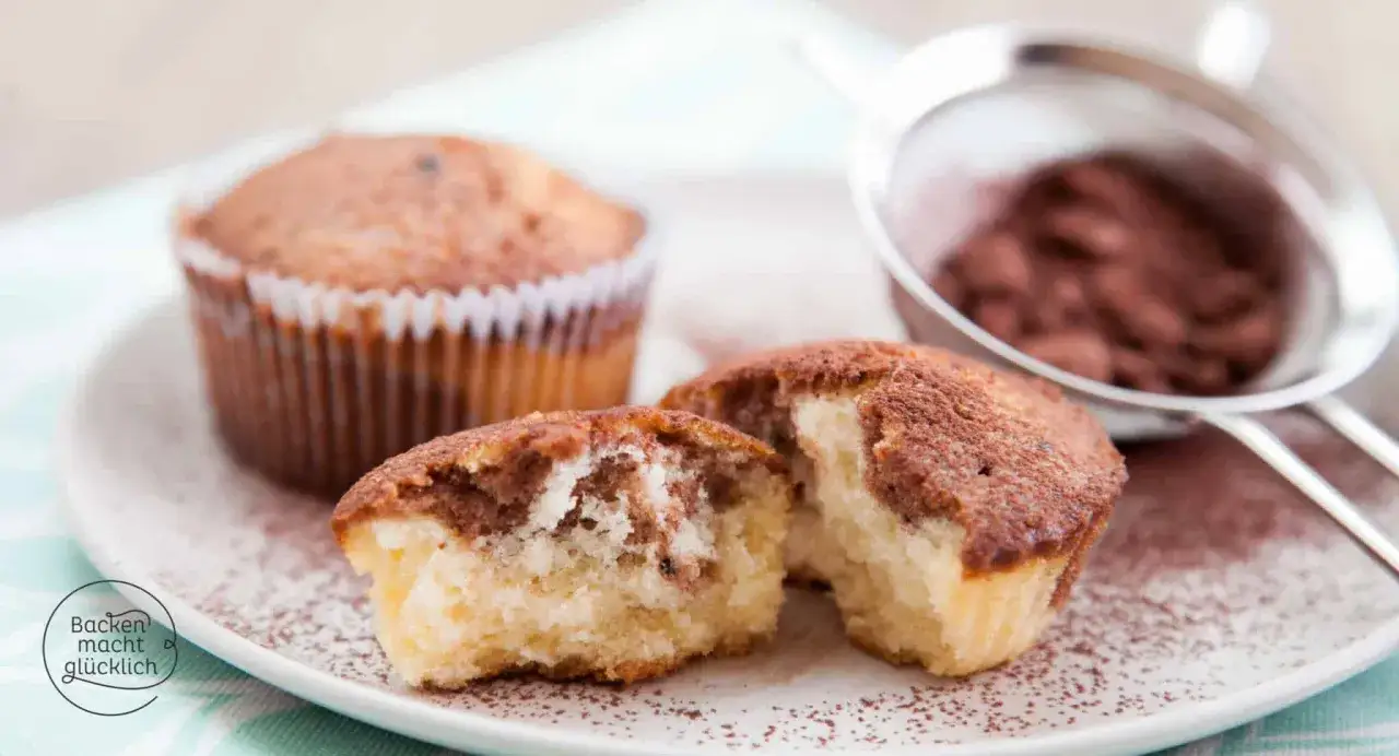 Zwei Marmor Muffins, einer angeschnitten, zeigen das sch&ouml;ne Muster. Ein Sieb mit Kakaopulver im Hintergrund. Perfekt f&uuml;r ein Marmor Muffins Rezept.