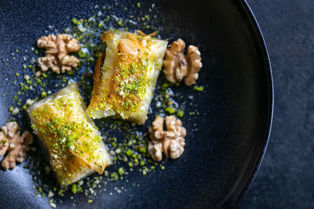 Zwei goldbraune Baklava-Stücke, bestreut mit gehackten Pistazien und Walnüssen, auf einem dunklen Teller. Ein köstliches Baklava Rezept.