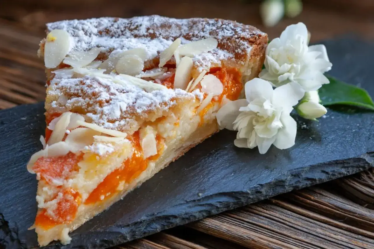 Ein St&uuml;ck weltbester Aprikosenkuchen, best&auml;ubt mit Puderzucker und Mandelbl&auml;ttchen, daneben zarte wei&szlig;e Bl&uuml;ten.