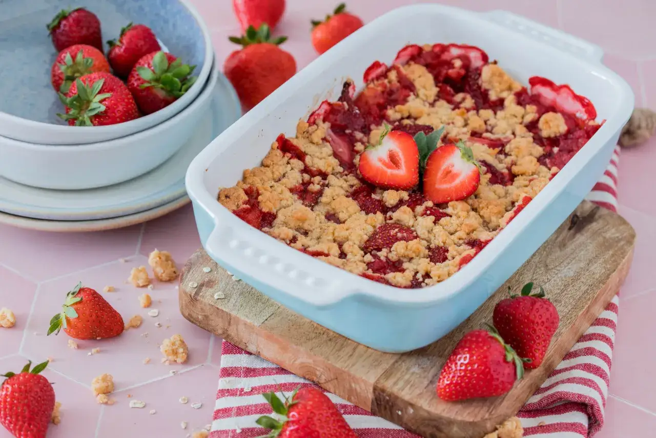 Ein frisch gebackener Erdbeer Crumble in einer hellblauen Auflaufform, garniert mit Erdbeerh&auml;lften. Frische Erdbeeren liegen daneben.