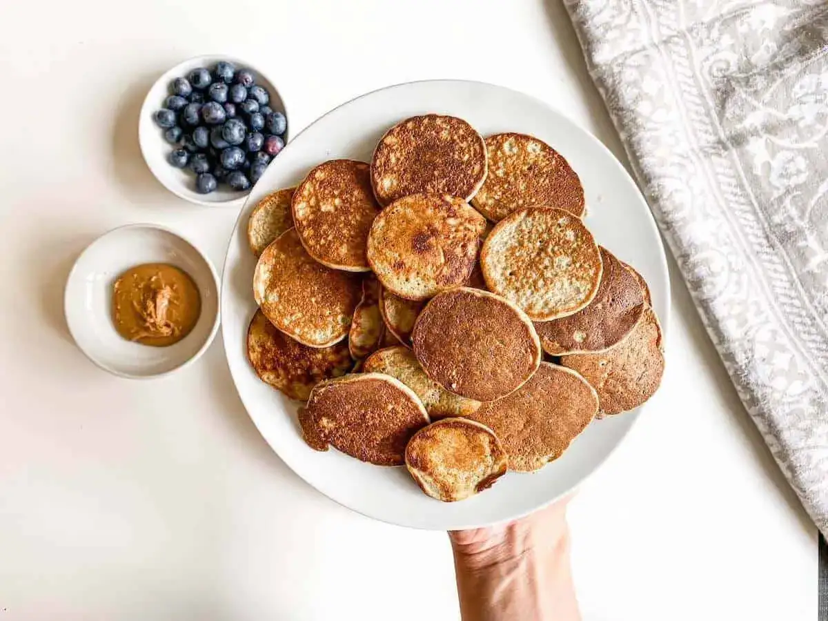Ein Teller voller kleiner, goldbrauner Baby Pancakes, daneben eine Sch&uuml;ssel mit Blaubeeren und Erdnussbutter. Perfekt f&uuml;r einen s&uuml;&szlig;en Start in den Tag!
