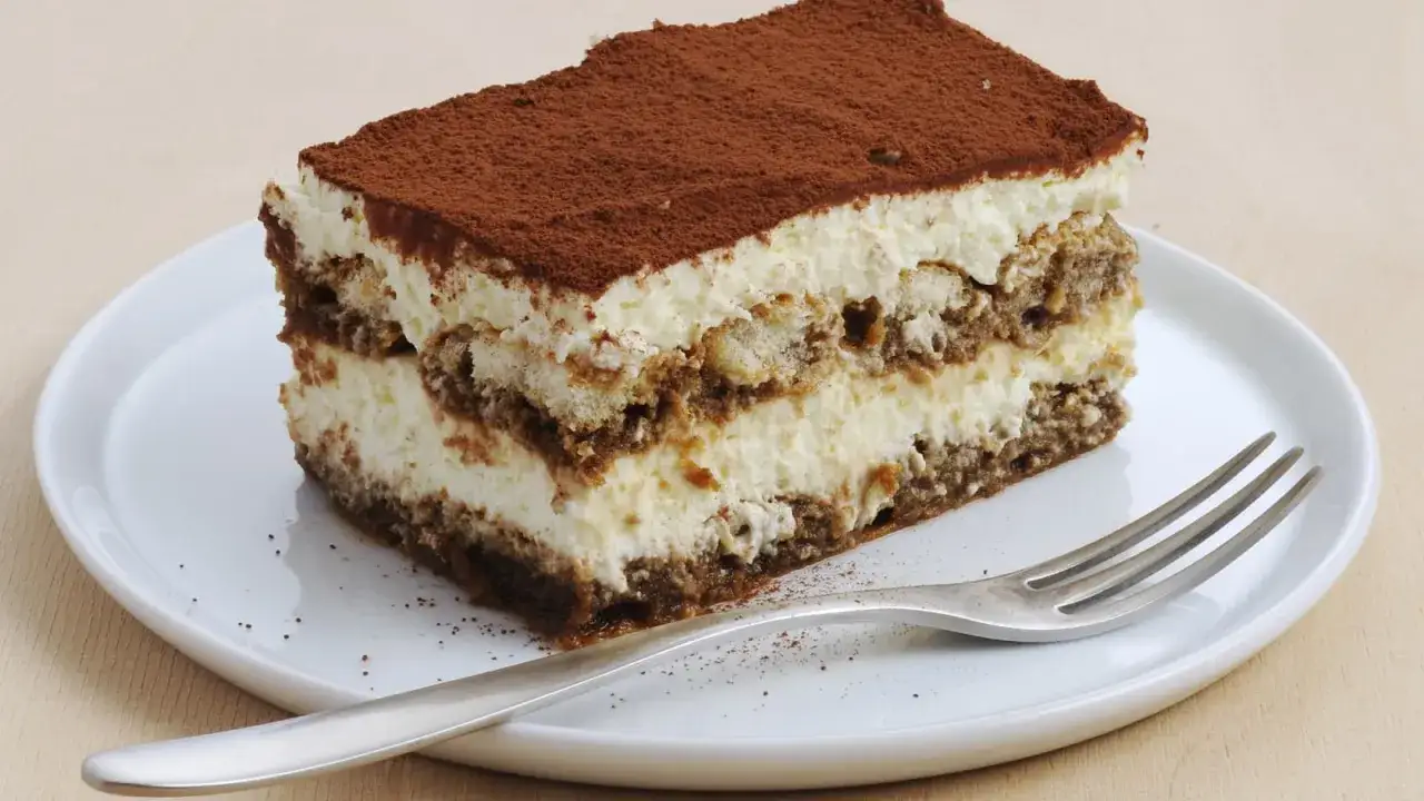 Ein St&uuml;ck cremiges Tiramisu ohne Ei, best&auml;ubt mit Kakao, auf einem wei&szlig;en Teller mit einer Gabel.