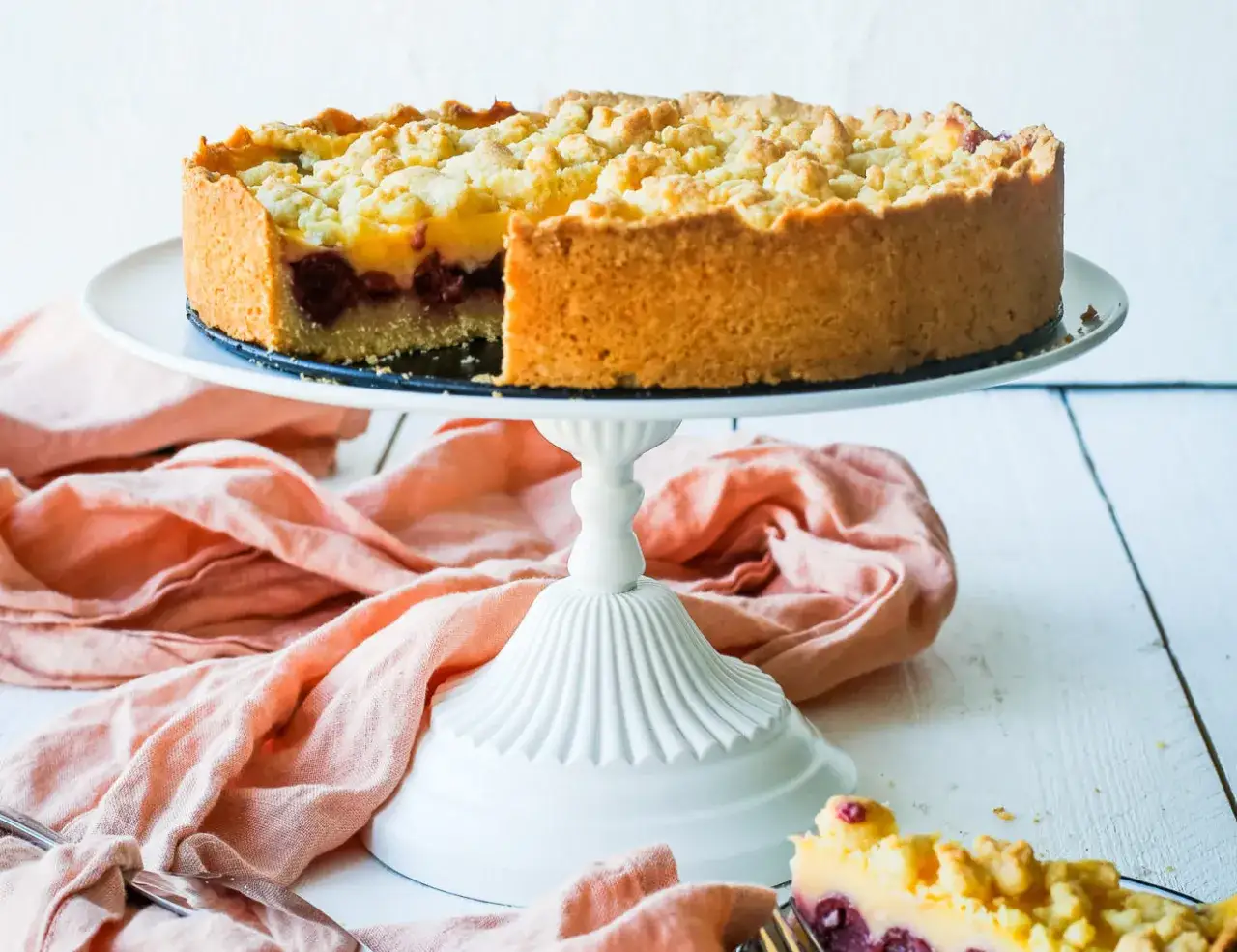 Ein k&ouml;stlicher Kirschkuchen mit Pudding und Streuseln auf einem wei&szlig;en Tortenst&auml;nder, daneben ein St&uuml;ck Kuchen.