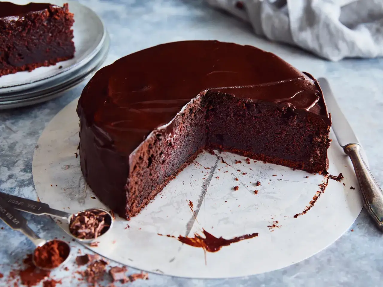 Ein saftiger schoko kuchen mit glänzender Glasur, ein Stück fehlt. Daneben liegen Löffel mit Kakaopulver und Schokostückchen.