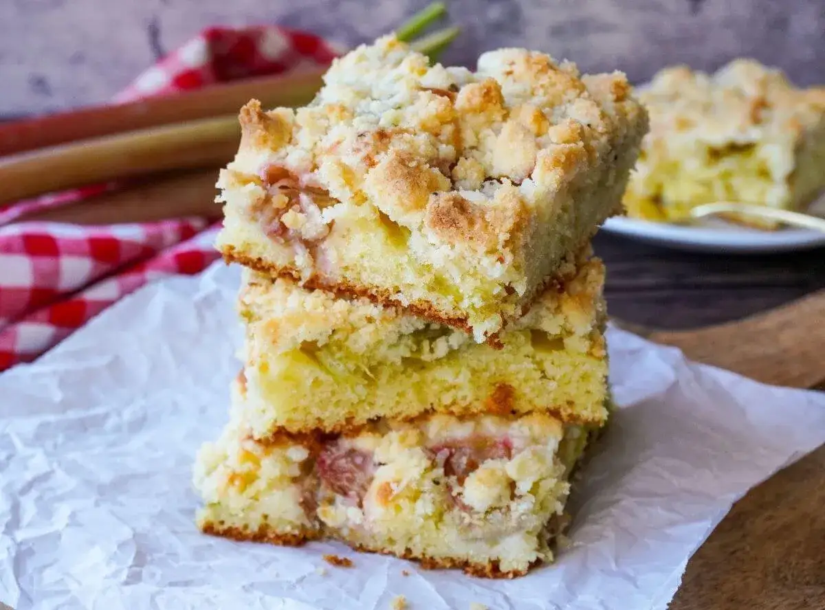 Saftiger Rhabarber Blechkuchen mit Streuseln, gestapelt auf wei&szlig;em Papier. Daneben liegen frische Rhabarberstangen.