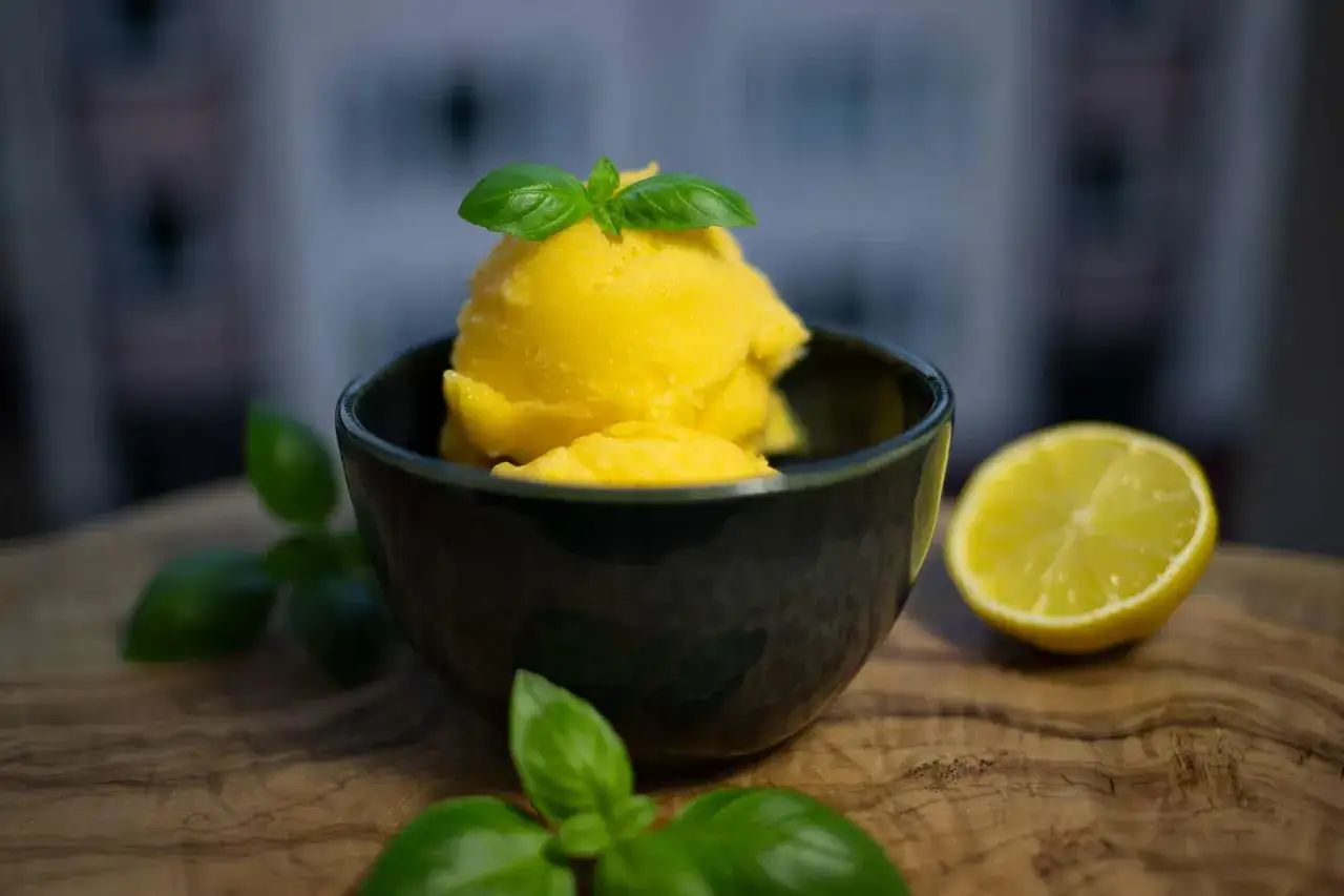 Ein Sch&auml;lchen mit leuchtend gelbem Mango Sorbet, garniert mit Basilikum. Daneben eine Zitronenh&auml;lfte.