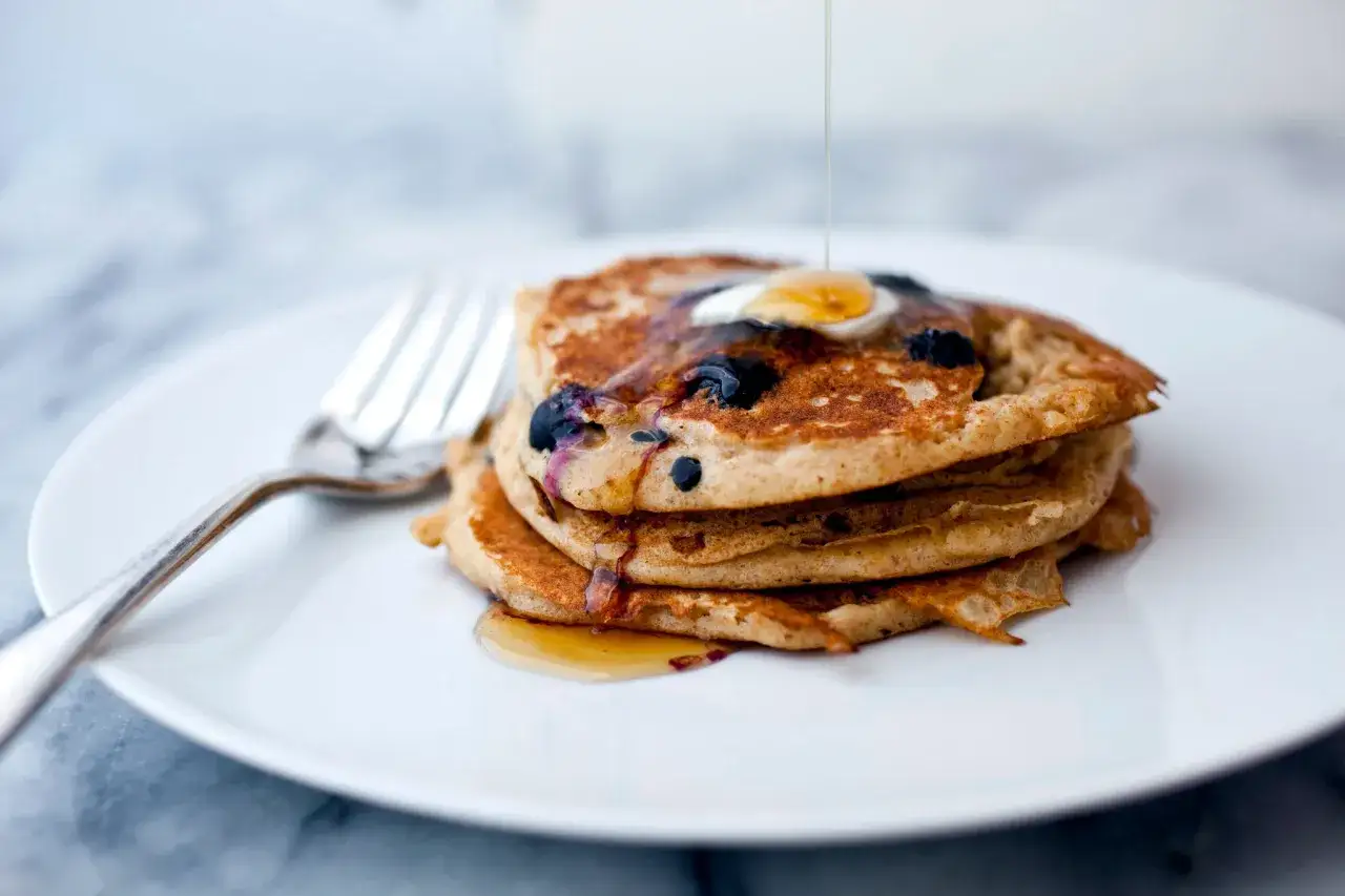 Ein Stapel gesunde Pancakes mit Blaubeeren, beträufelt mit Sirup und einem Klecks Butter. Daneben liegt eine Gabel.