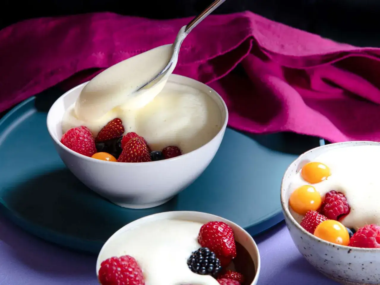 Cremige Panna Cotta mit frischen Beeren und Physalis, serviert in weißen Schalen. Ein Löffel hebt die luftige Masse an.