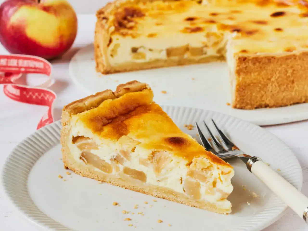 Ein Stück saftiger Apfelkuchen mit Quark auf einem Teller, daneben ein ganzer Kuchen und ein Apfel.