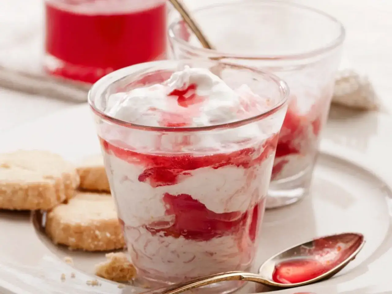 Cremiges Dessert mit Rhabarberkompott und Sahne, serviert mit Keksen. Ein Löffel mit roter Sauce liegt daneben.