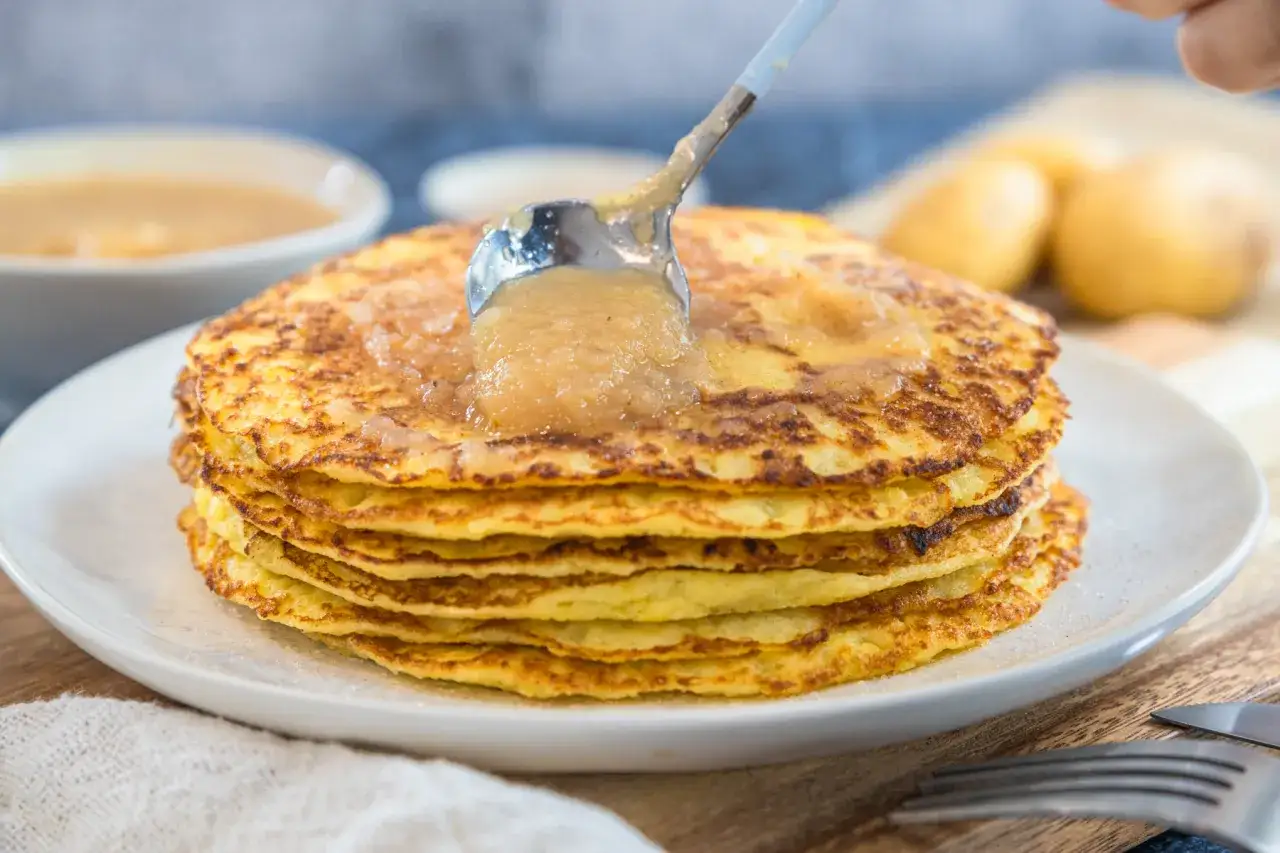 Ein Stapel goldbrauner Kartoffelpuffer, zubereitet im Thermomix, wird mit Apfelmus betr&auml;ufelt.