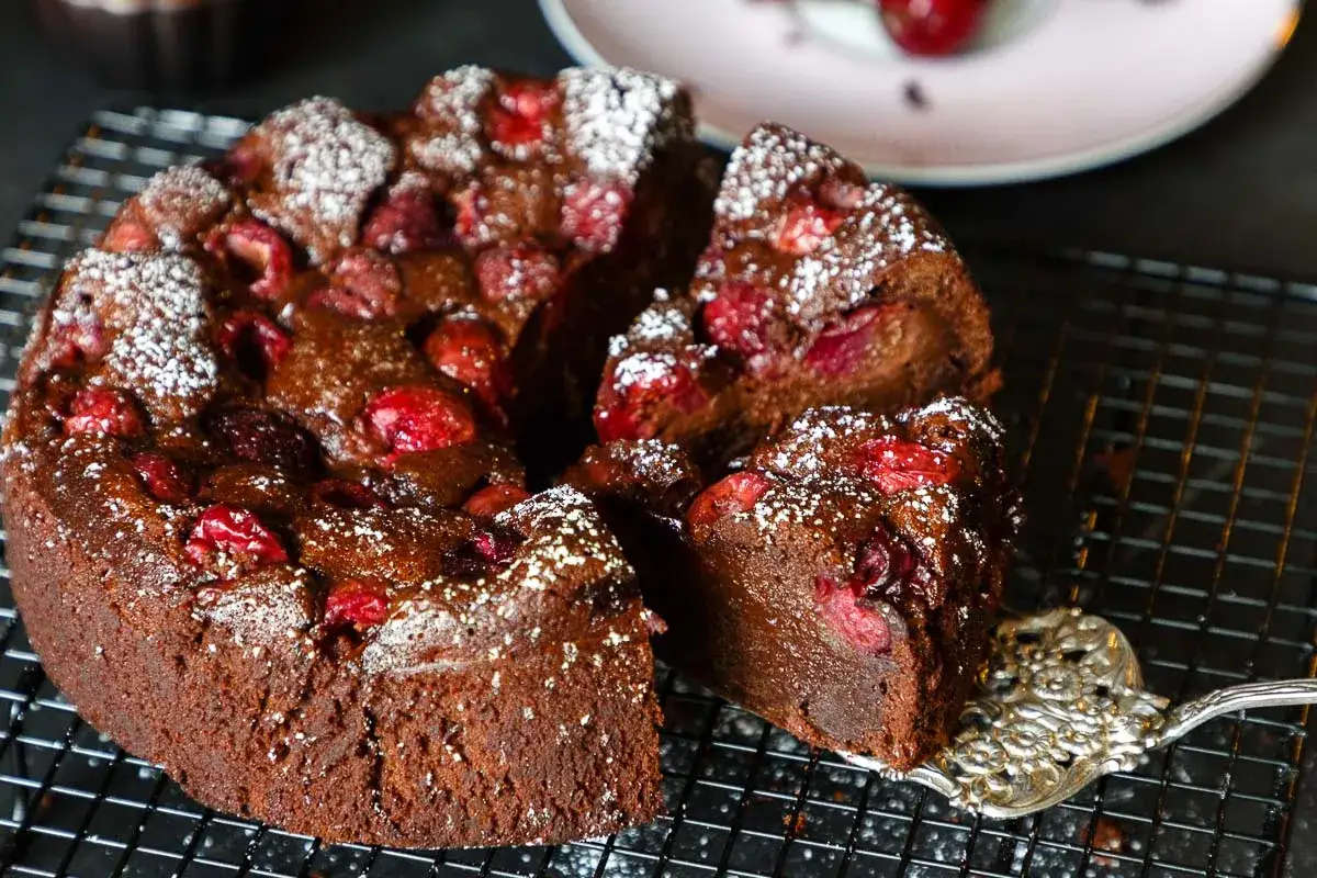 Ein saftiger schoko kirsch kuchen, bestäubt mit Puderzucker, wird auf einem Gitter präsentiert. Eine Tortenspitze hebt ein Stück heraus.