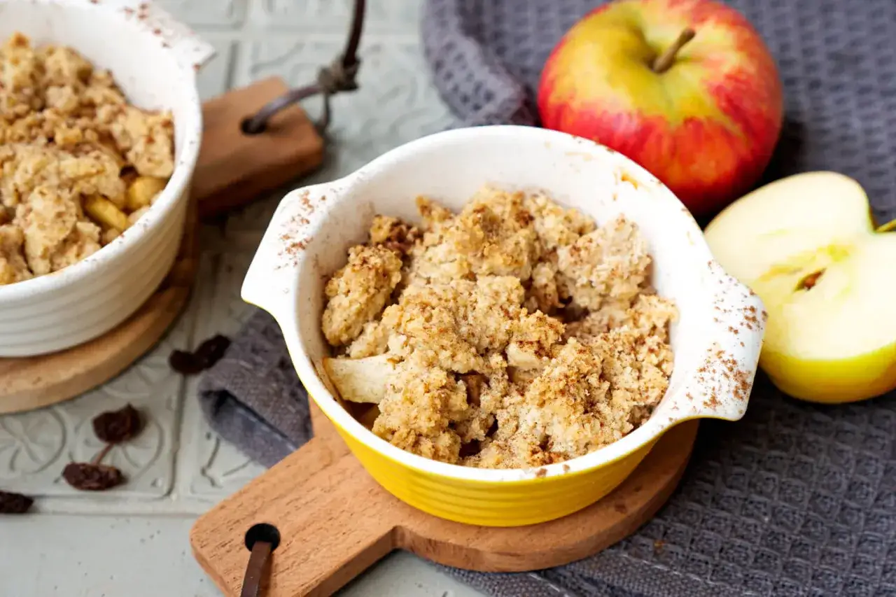 Zwei Schalen mit warmem Apfel-Crumble, daneben ein ganzer und ein halber Apfel. Zimt bestäubt die knusprige Krume.
