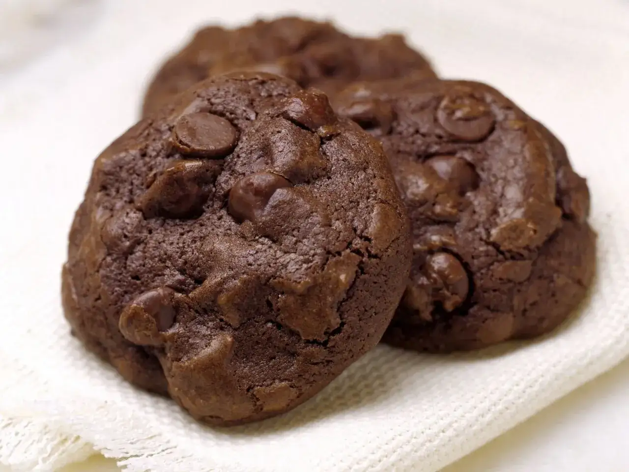 Drei saftige schoko cookies mit vielen Schokost&uuml;ckchen liegen auf einem wei&szlig;en Tuch.