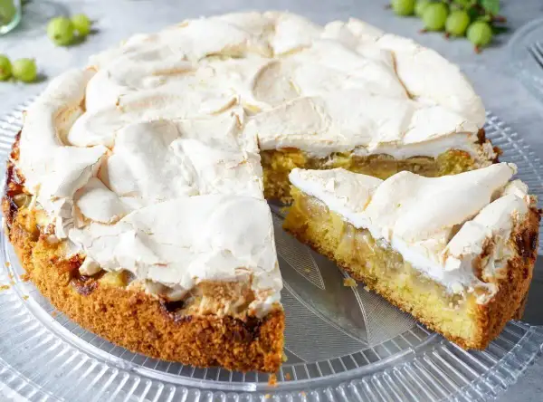 Omas stachelbeerkuchen mit luftiger Baiserhaube auf einem Tortenteller. Ein Stück ist bereits angeschnitten.