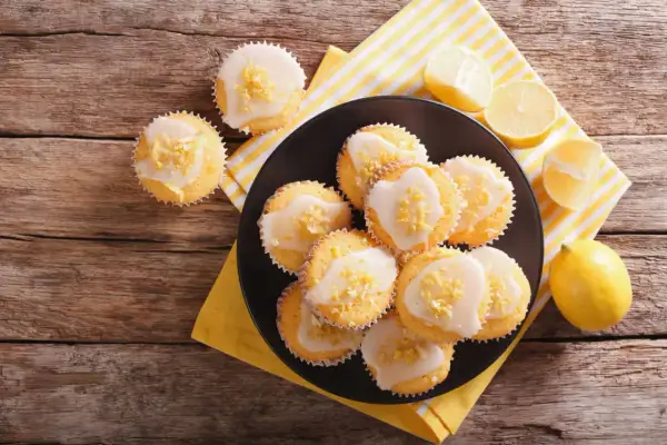 Zitronen-Muffins, frisch aus dem Thermomix, mit Zuckerguss und Zitronenschale bestreut, auf einem Holztisch.